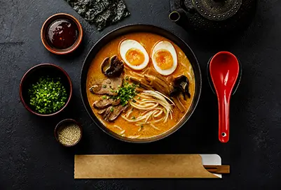 Bowl of ramen with different veggies, meats, and eggs on a properly set table.