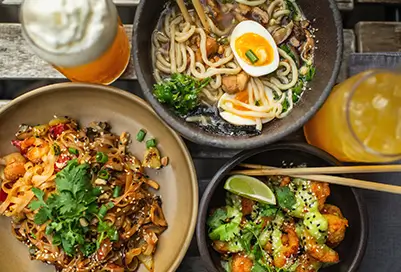 Different bowls of noodles and curries on table with beer and tea.